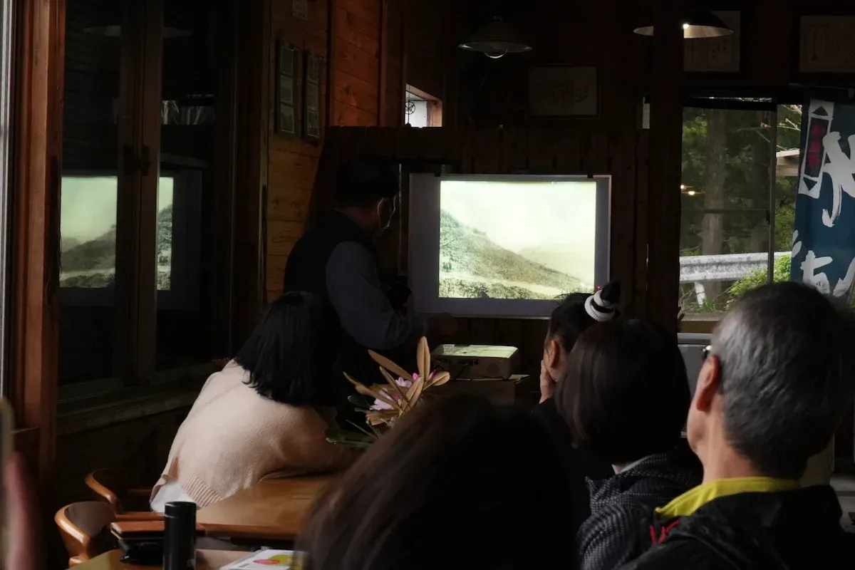 The scene shows Isao Yamada explaining using a projector, with participants listening.